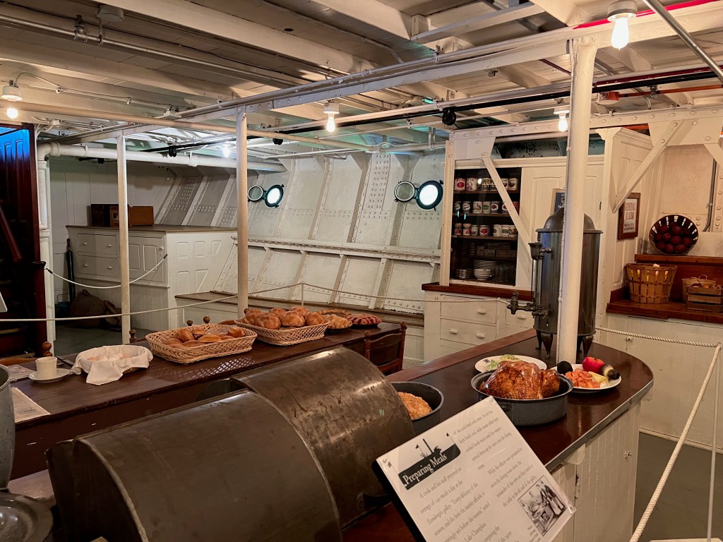 Interior of a historical ship's galley, featuring a wooden counter with food items such as bread and fruits, cabinets with canned goods in the background, and portholes providing natural light.