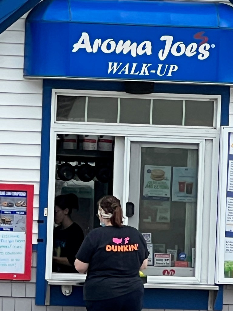 A person wearing a black shirt with 'DUNKIN'' on the back stands at a walk-up window labeled 'Aroma Joes'. A menu board is visible on the left.