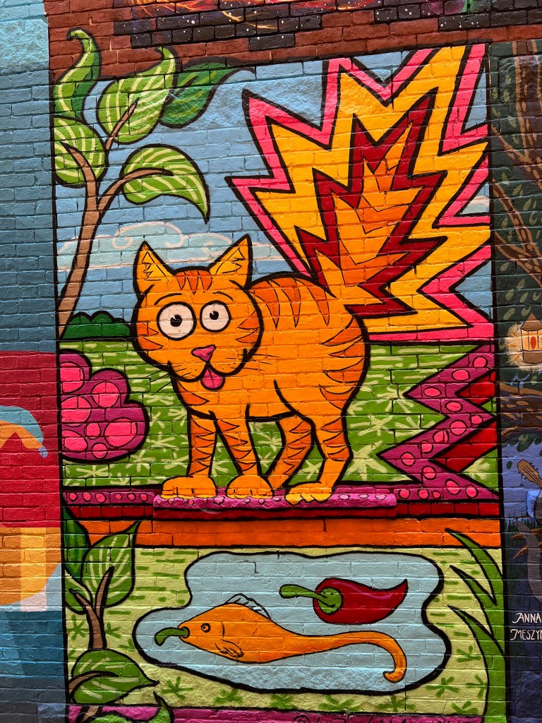 A colorful mural featuring a cartoonish orange cat with large eyes and a playful expression, standing on a bright green background with pink flowers and abstract shapes. Below the cat, there is a whimsical depiction of a fish and a red chili pepper.