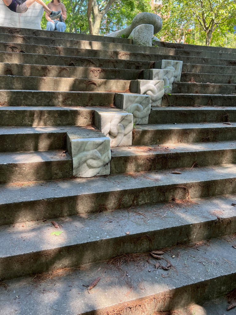Stairs in a park featuring sculpted stone blocks with intricate designs, surrounded by trees and sunlight.