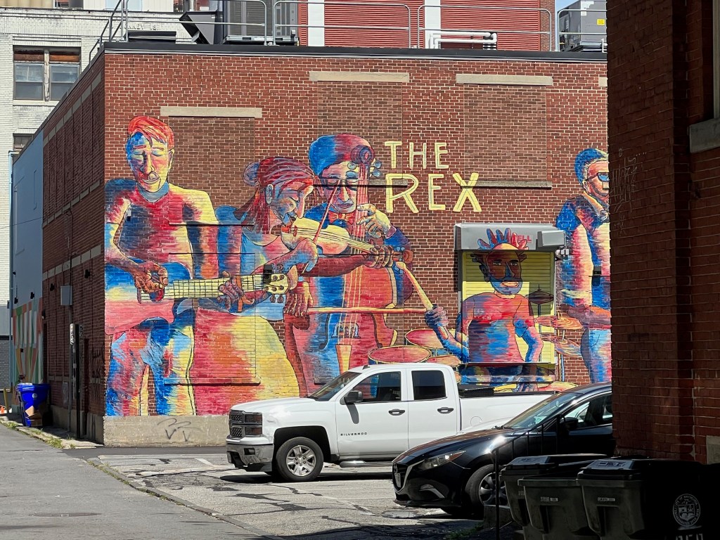 A vibrant mural depicting musicians playing various instruments on a brick wall. The mural features colorful figures of a guitarist, violinist, and drummer, with the name 'THE REX' prominently displayed above.