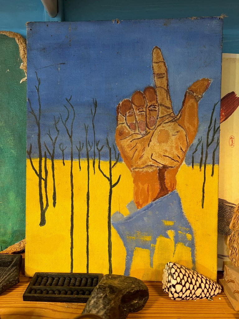 A painting depicting a raised hand against a blue sky with bare trees in the background and a yellow field in the foreground.