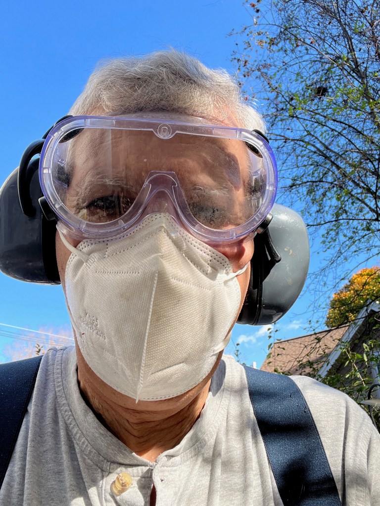 An older adult wearing goggles and a mask, with headphones visible, outdoors on a sunny day.
