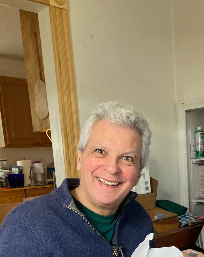 A man with gray hair smiling broadly while sitting at a table in a kitchen setting, wearing a blue sweater, with various kitchen items visible in the background.