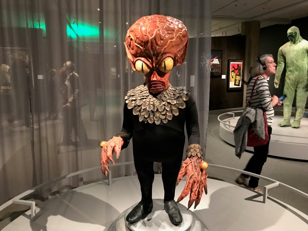 A life-sized alien creature display from a movie, featuring an oversized head, yellow eyes, and claw-like hands, standing on a circular pedestal in a museum setting.