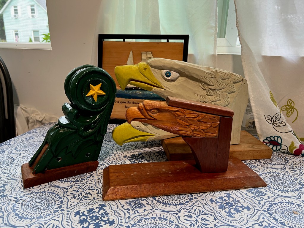 Three carved wooden figureheads displayed on a table, featuring an eagle head, a stylized figure, and a detailed bird head, with a blue and white patterned tablecloth in the background.
