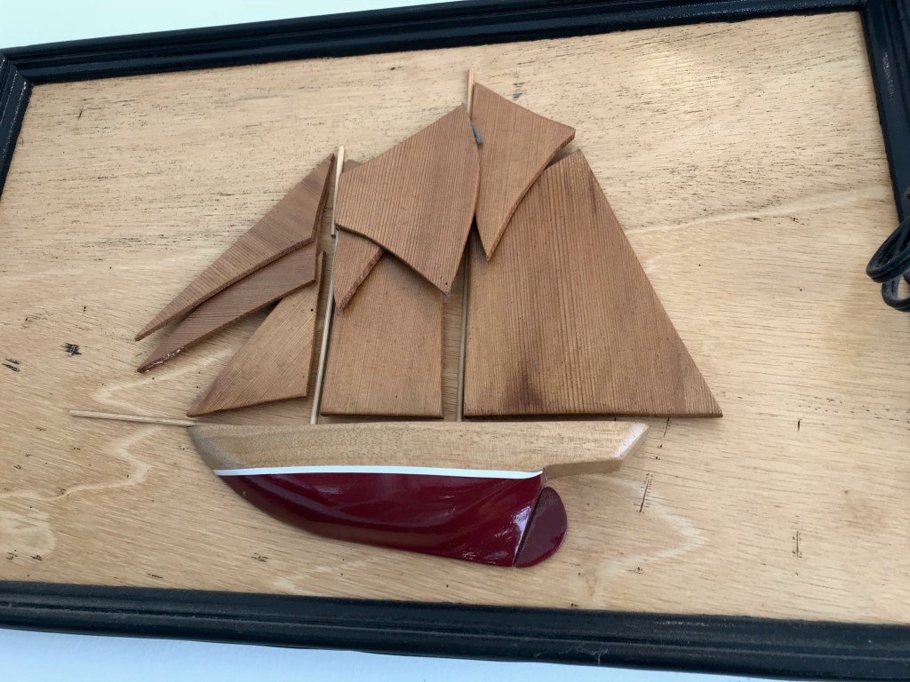 A wooden wall art piece depicting a sailboat with three sails and a red hull, mounted on a natural wood background.