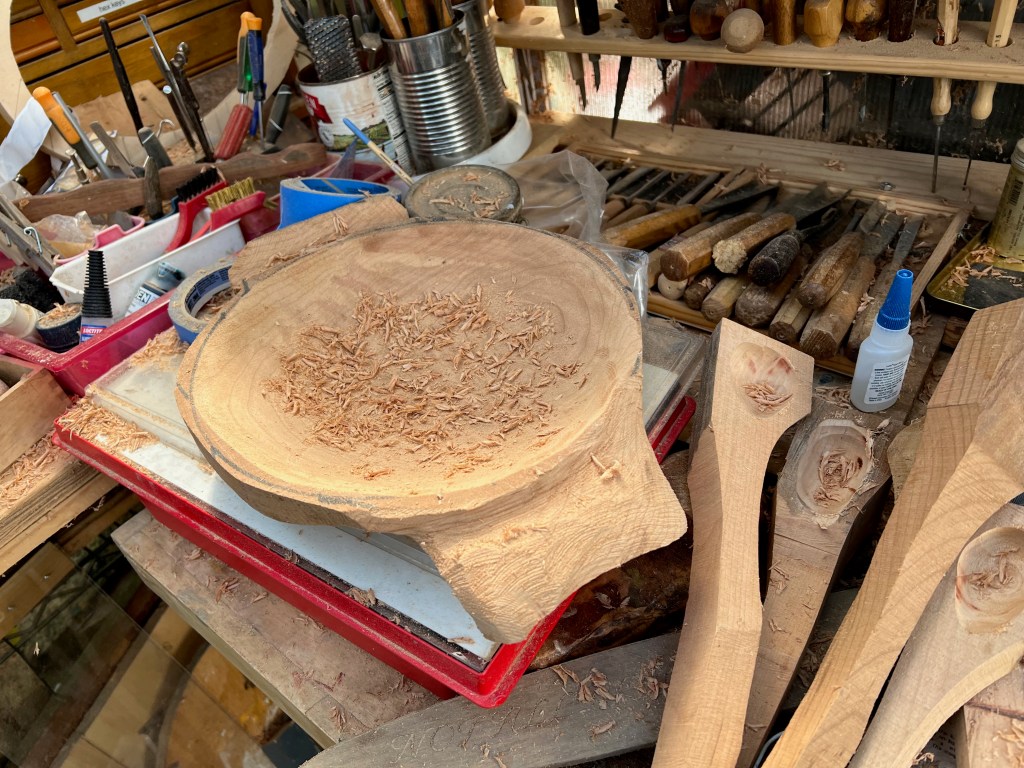 A woodworking workspace featuring a round wooden bowl with shavings scattered on a work surface. Various woodworking tools and materials are organized around the bowl.