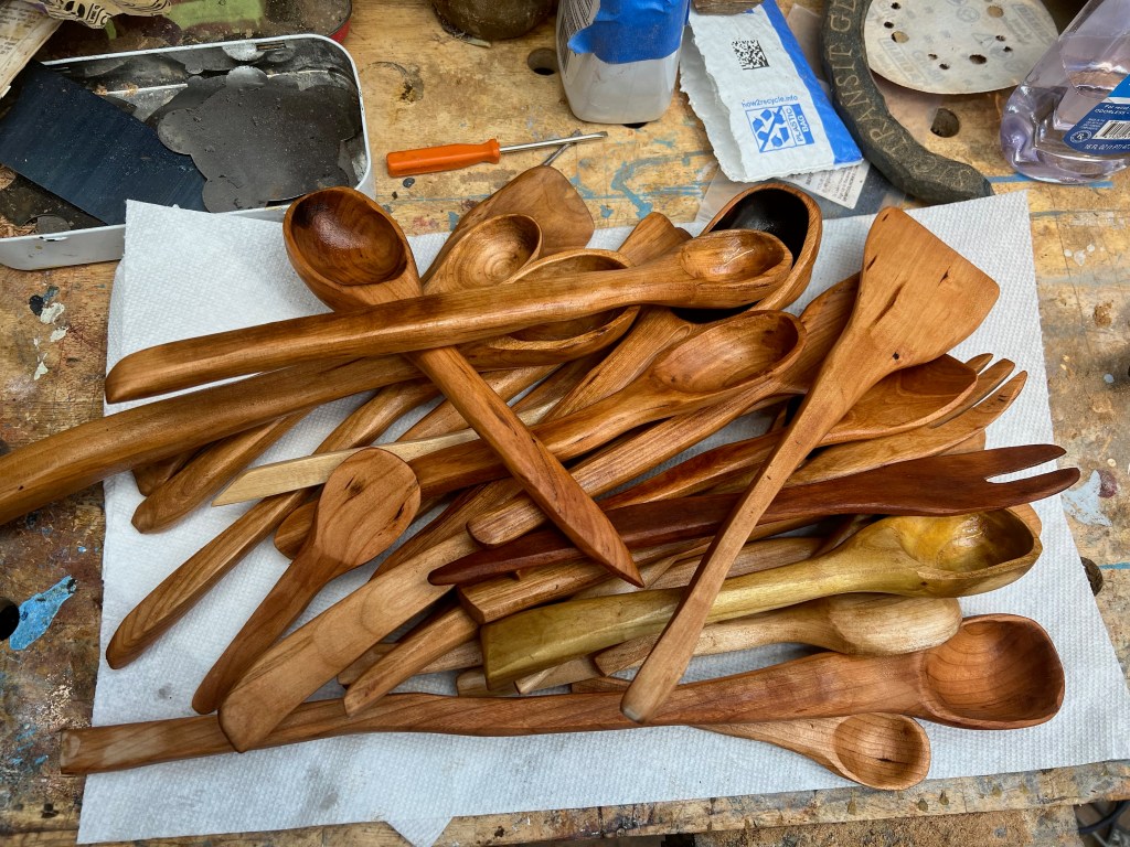 A collection of handcrafted wooden spoons and utensils arranged on a white paper towel, with a workbench background showing other tools and materials.