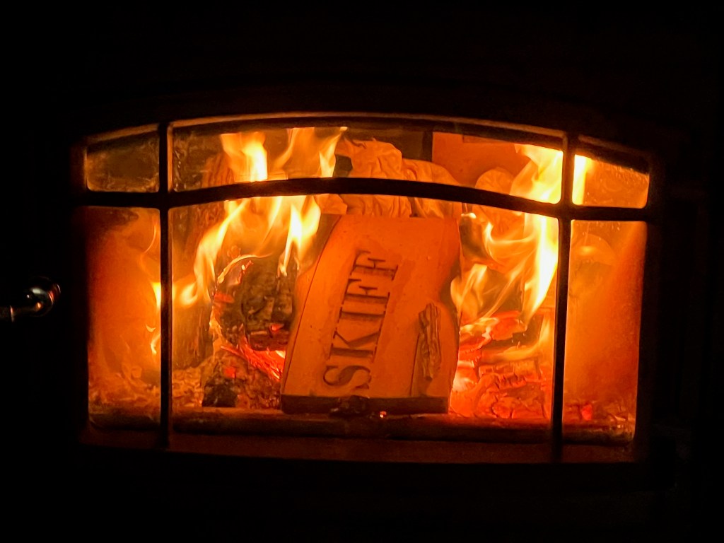 A close-up view of a fireplace with flames flickering and logs burning inside it, with a visible piece of wood labeled 'SKIFE'.