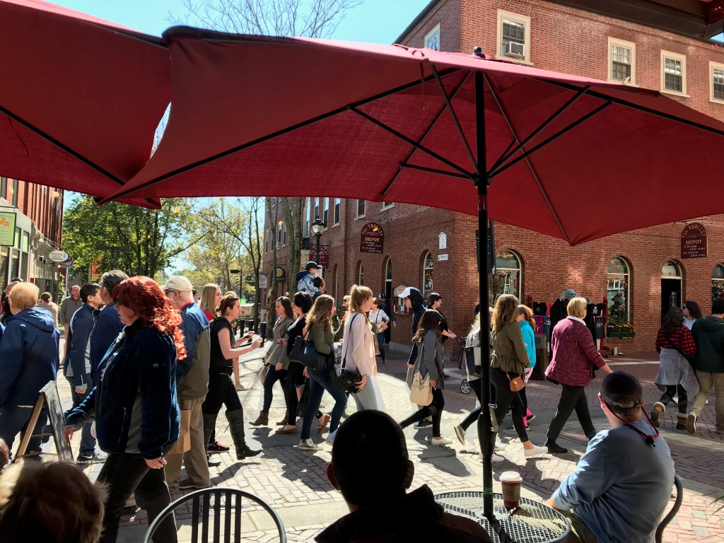 A bustling street scene with people walking under red umbrellas, brick buildings in the background, and shops lining the sidewalks.
