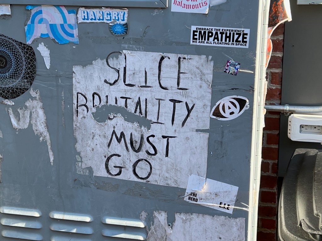 Graffiti on a wall reading 'SILENCE BRUTALITY MUST GO' with various stickers and peeling paint.