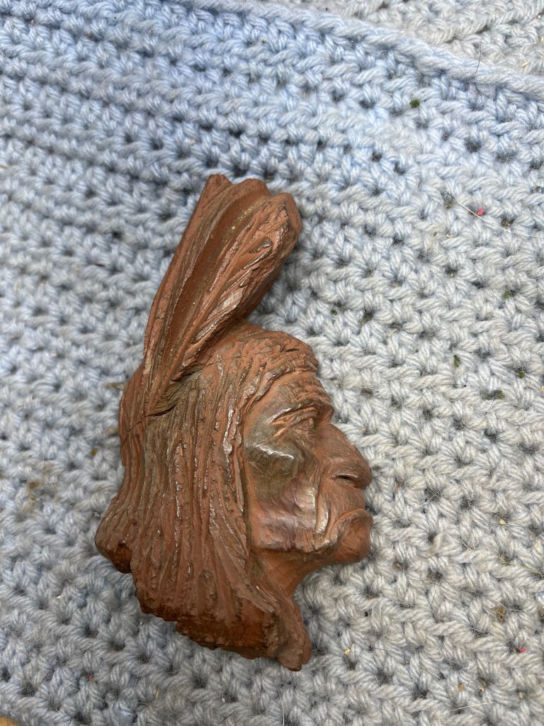 Side profile of a carved clay figure resembling a Native American chief, featuring long hair and a feather headdress, placed on a textured gray background.