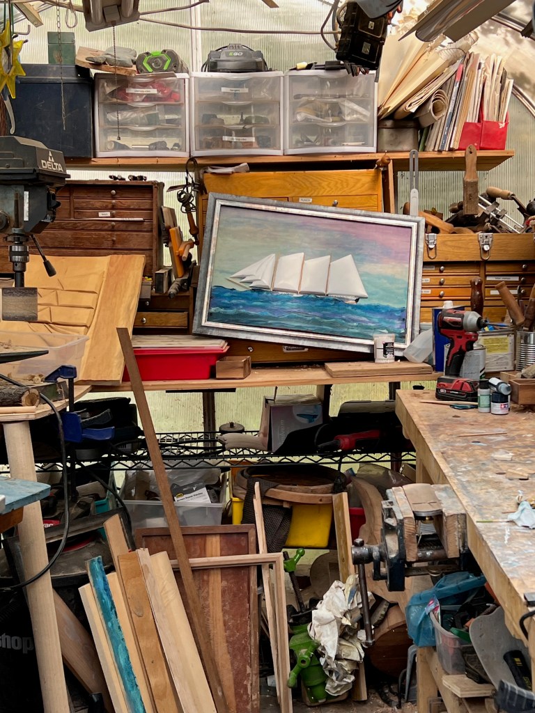 A cluttered workshop featuring various wooden pieces, tools, and storage boxes, with a painted seascape of sailing ships prominently displayed on the wall.