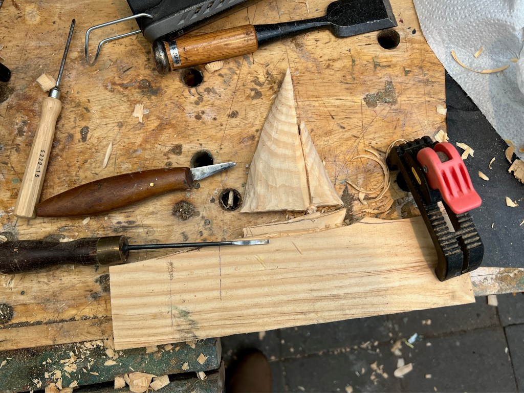 A wooden workbench with various carving tools, including a chisel, a knife, and a small triangular wood piece, surrounded by wood shavings.