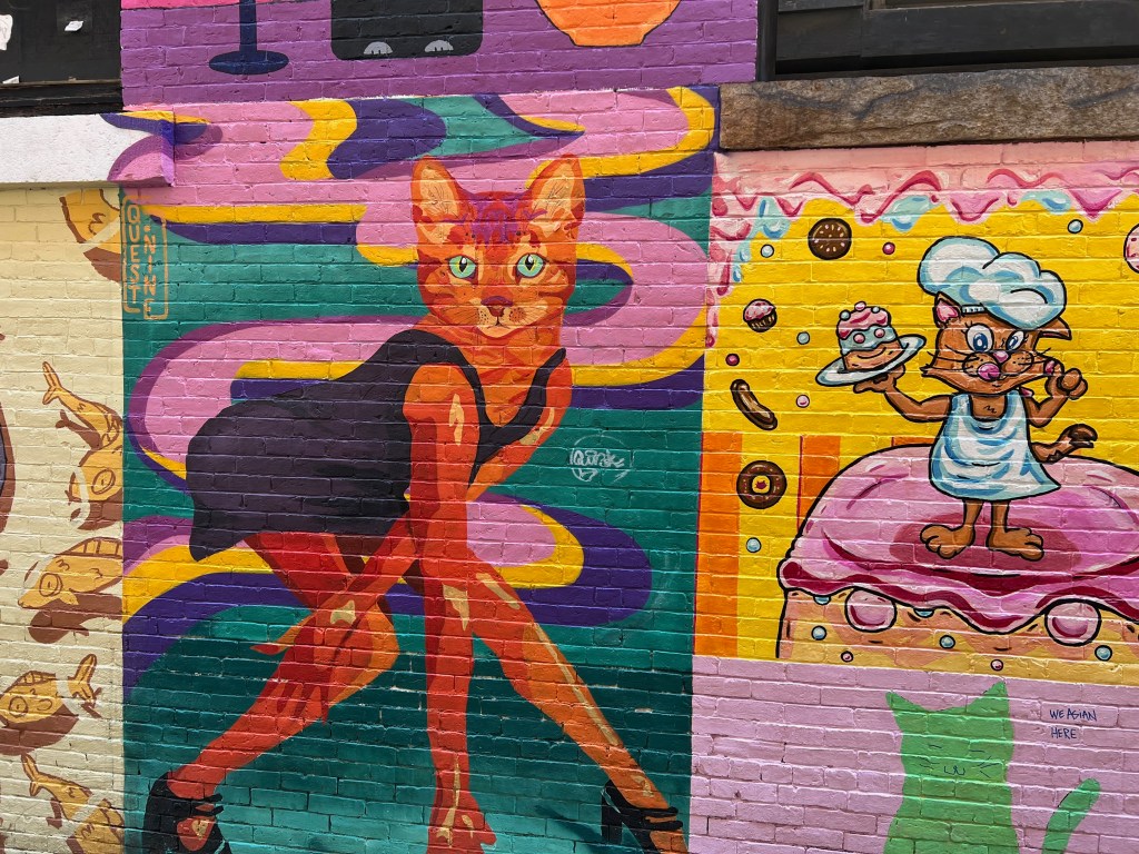 Colorful mural featuring a cat in a black dress striking a pose, with abstract shapes and other whimsical characters in the background.
