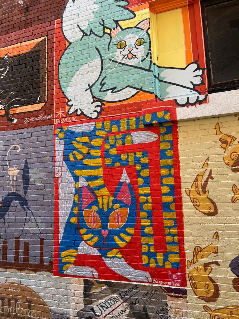 Colorful mural of cats on a brick wall featuring a playful green cat and a stylized blue and yellow cat design.