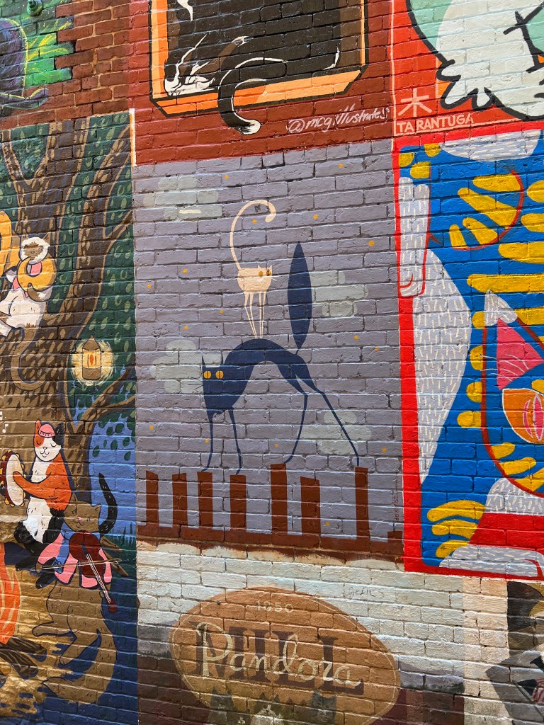 A colorful mural depicting various cats and whimsical scenes, including a cat with a crown and other artistic representations on a brick wall.