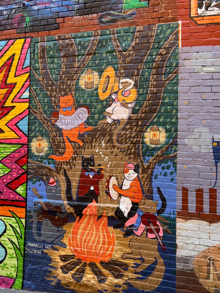 A colorful mural depicting cats around a campfire, playing musical instruments, set against a brick wall.