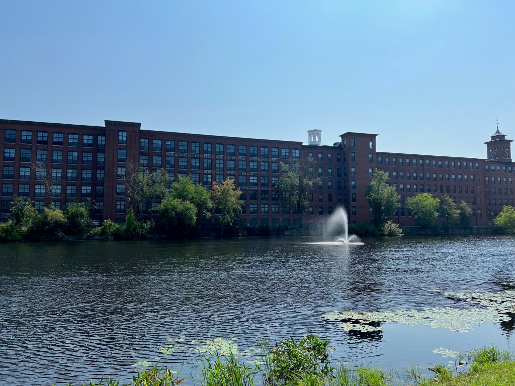 Historic industrial building along a river, featuring a fountain and surrounding greenery under a clear blue sky.