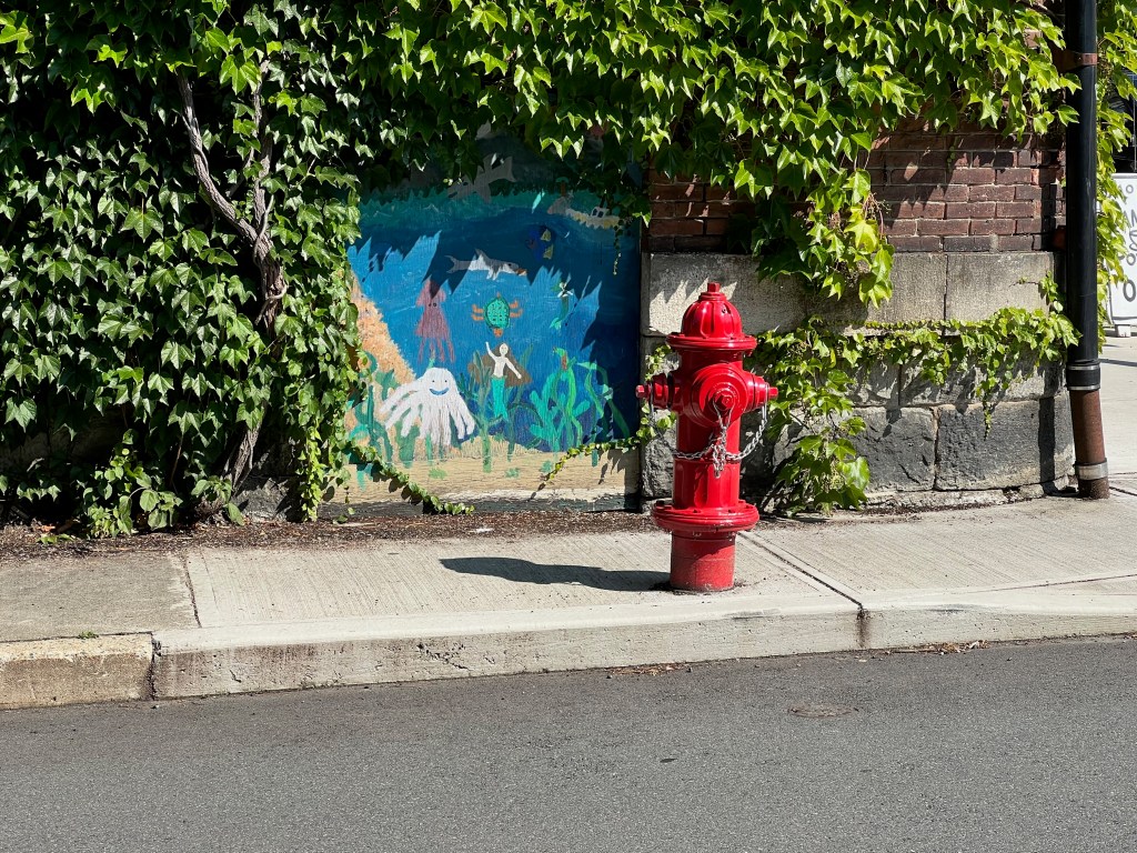 A bright red fire hydrant beside a wall covered in ivy, accompanied by a colorful mural depicting underwater scenes with various sea creatures.