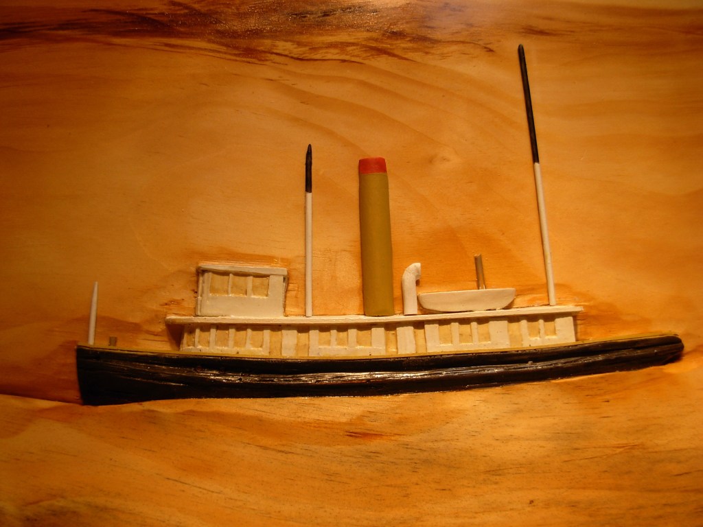 A detailed model of a steamboat displayed against a wooden background, featuring a black hull, white superstructure, and distinctive smokestack.