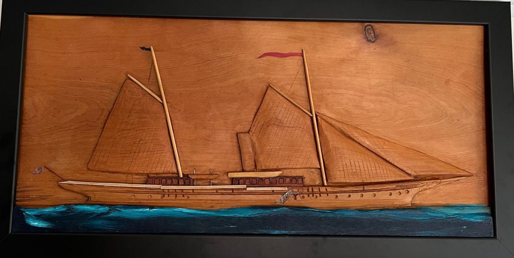A carved wooden artwork depicting a traditional sailing ship, showcasing two large sails and intricate details, with a blue water background.