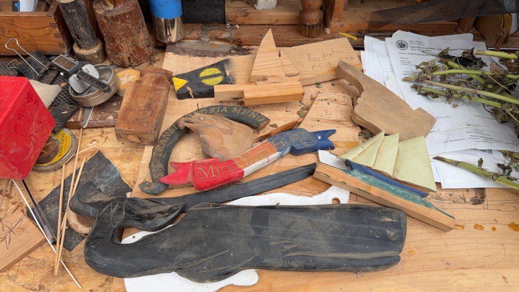 A cluttered workbench filled with various carved wooden pieces, tools, and materials, including a red box, wooden blocks, and scattered papers.