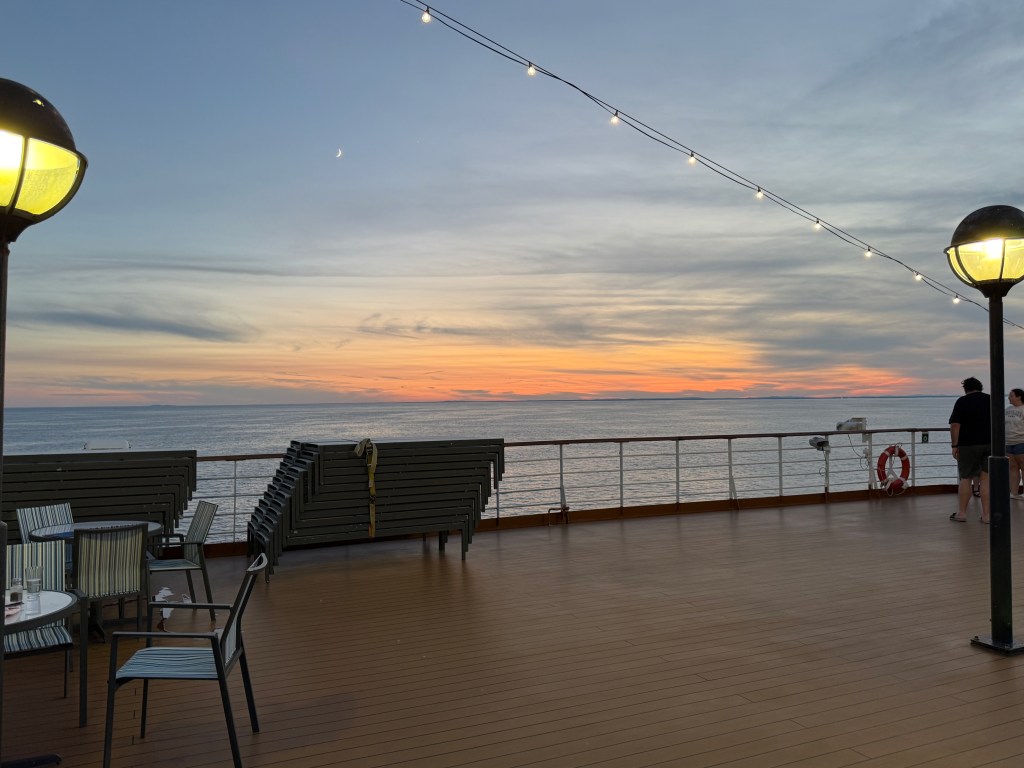 View of a deck on a ship during sunset, with a calm ocean and a colorful sky. Two people walking in the background, tables and chairs in the foreground, and decorative lights overhead.