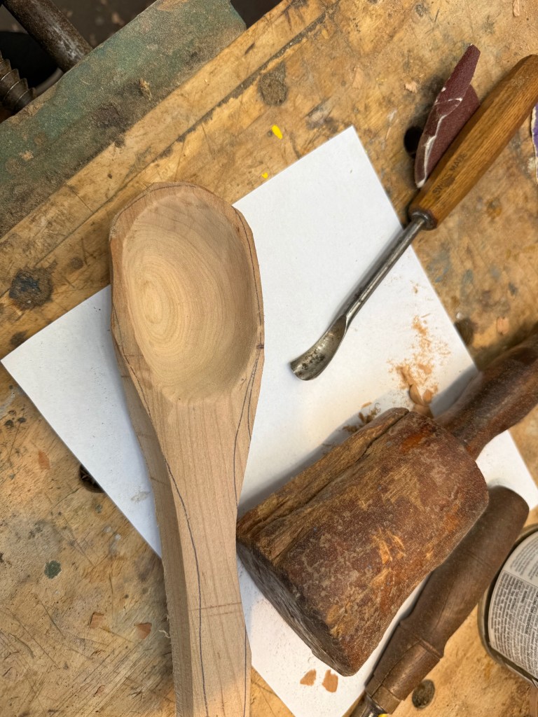 A wooden spoon in the process of being carved, lying on a workbench with a sheet of paper, a wooden mallet, and carving tools.
