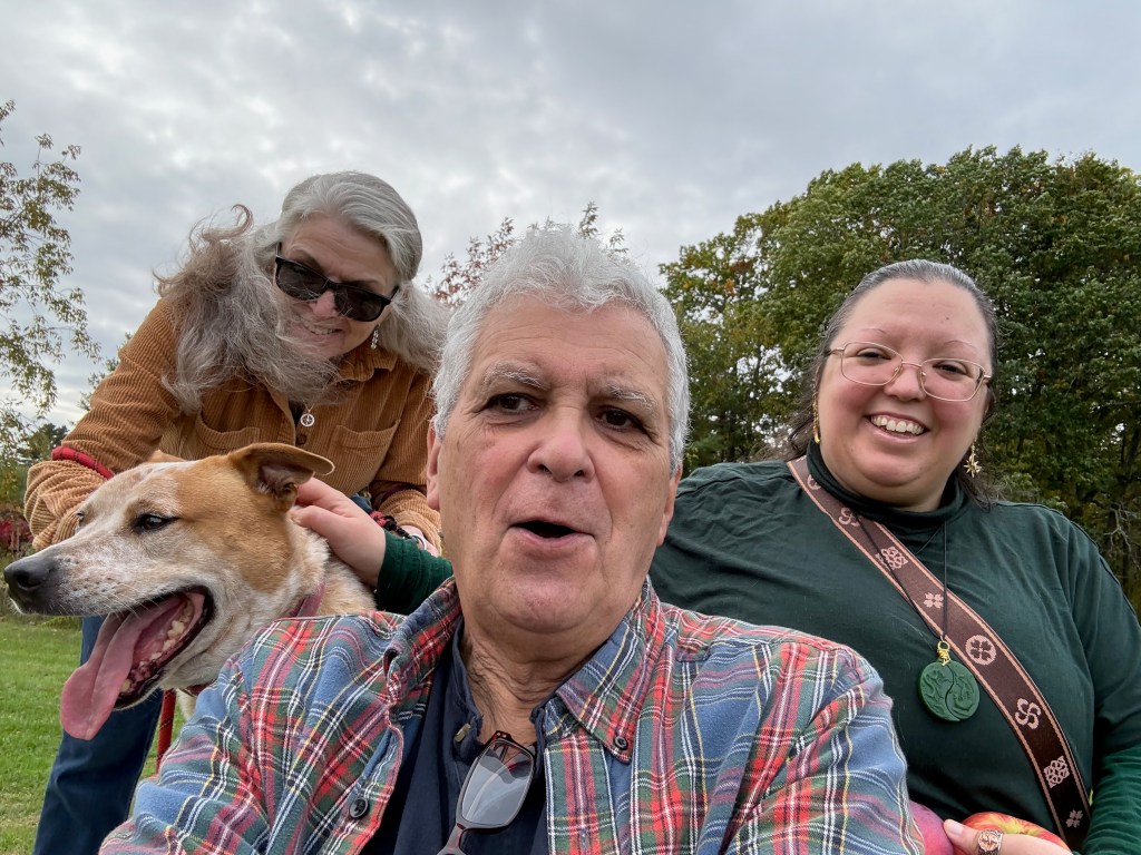 A group selfie featuring three people smiling outdoors, with a dog in the foreground. The background includes trees and a cloudy sky.