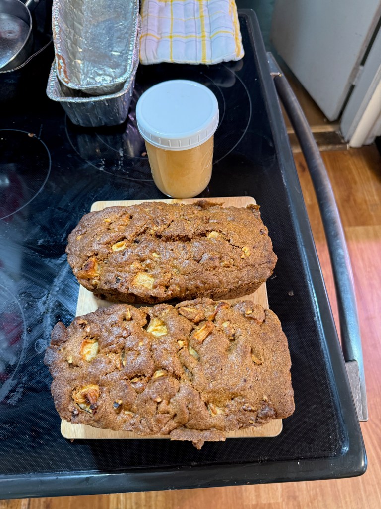 Two loaves of baked apple bread on a wooden cutting board, with a jar of peanut butter beside them. The background features a stovetop with cooking utensils and a colorful dish towel.